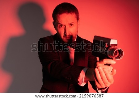 Man in an elegant suit handling a Super 8 camera in classic secret agent pose, shooting at camera  Red light illumination 