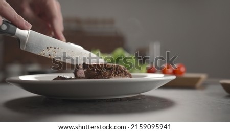 Man slicing beef steak on white plate on kitchen countertop, wide photo