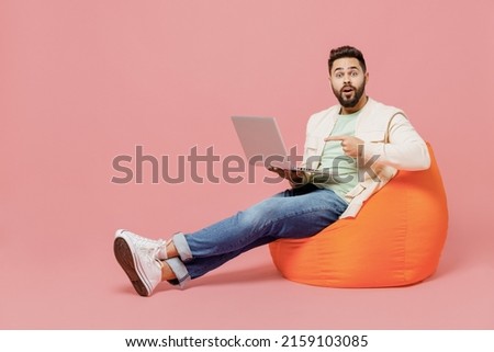 Full body young fun happy man in trendy jacket shirt sit in bag chair hold use work point finger on laptop pc computer isolated on plain pastel light pink background studio. People lifestyle concept