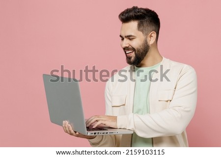 Young smiling happy cheerful caucasian man 20s wearing trendy jacket shirt hold use work on laptop pc computer isolated on plain pastel light pink background studio portrait. People lifestyle concept