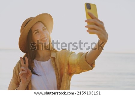 Sunlit young woman in straw hat shirt summer casual clothes do selfie shot on mobile cell phone show v-sign outdoors at sunrise sun dawn over sea background People vacation lifestyle journey concept
