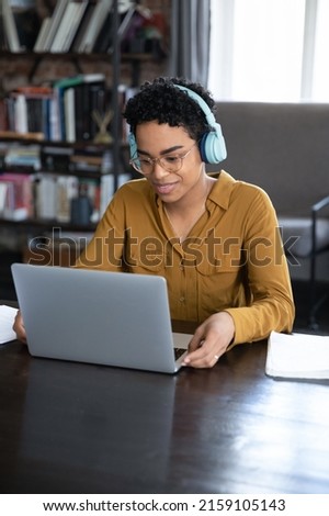 Young African woman sit at desk with laptop listen audio course through wireless headphones, take part in video call by business or study with online tutor, gain new knowledge use modern tech concept Royalty-Free Stock Photo #2159105143