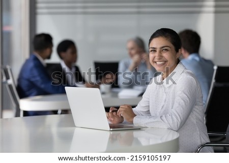 Indian businesswoman satisfied by company position, career growth at desk with laptop smile pose look at camera, diverse colleagues on background. Successful promoted company employee portrait concept Royalty-Free Stock Photo #2159105197