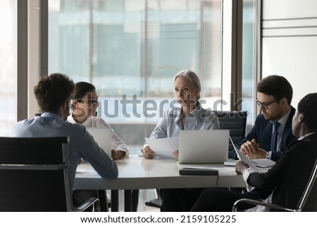 Older female executive manager teach diverse staff, take part in group meeting in office boardroom, analyze, make paperwork review, develop marketing plan, explain new strategy to workers at briefing Royalty-Free Stock Photo #2159105225