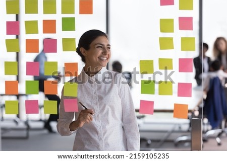 Indian woman office employee create visual plan for colleagues, prepare for briefing, develop strategy, make notes use colorful sticky notes attached on glass wall smile looks aside. Workflow concept