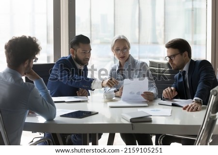 Serious multiracial older and young businesspeople gathered in boardroom discuss financial statistics, analyze sales report, forecasting work together at office meeting. Teamwork, negotiations concept Royalty-Free Stock Photo #2159105251