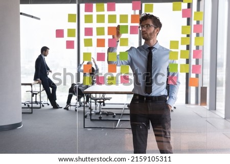 Serious businessman managing project tasks on sticky notes, writes start up business ideas using colorful post it stickers, plan corporate strategy on glass board. Creative priority to-do list concept Royalty-Free Stock Photo #2159105311