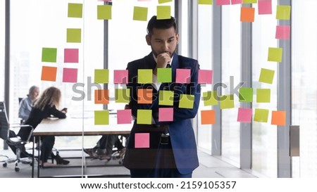 Young pensive Indian businessman in suit stand near glass wall staring at attached adhesive colored memory sticky post- reminders notes with visual tasks, business ideas, new start-up looks focused