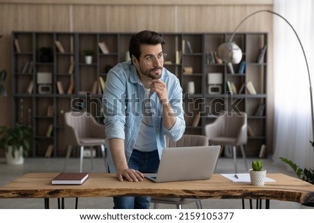 Work on idea. Thoughtful casual male in glasses stand by desk with pc look at distance touch chin create project ponder on business plan. Pensive young man scientist do research think on new solution Royalty-Free Stock Photo #2159105377