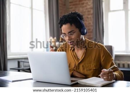 Serious diligent African woman student studying listen audio course through wireless headphones, take notes in copybook, makes assignment, improve foreign language knowledge use tech, e-learn concept Royalty-Free Stock Photo #2159105401