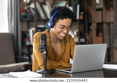 Smiling young African woman podcaster in headphones sit at desk in front of laptop and microphone. Share information, make audio podcast for internet audience, lead on-line stream, blogging concept Royalty-Free Stock Photo #2159105443