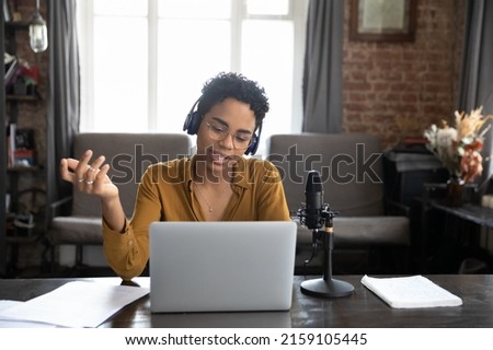 African woman wear wireless headphones sit at desk looks at laptop screen make speech talk into mike, records or lead live stream via internet in real time. Podcast, online radio program event concept Royalty-Free Stock Photo #2159105445