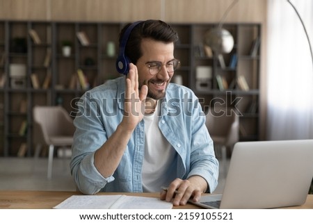 Got an answer. Young bearded male student in headset take part in online educational event raise hand up prepare to respond on teacher question. Polite millennial guy wave hand meet tutor by pc screen Royalty-Free Stock Photo #2159105521