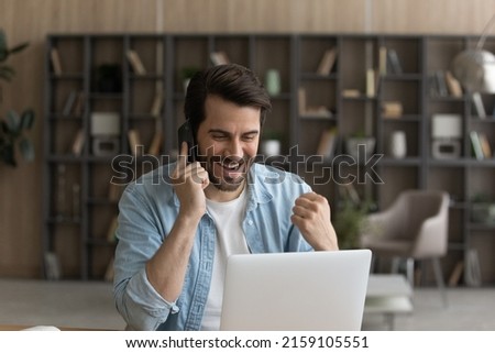Business win. Overjoyed millennial businessman working from home sit by laptop discuss perfect sales result on phone with colleague. Excited young man call friend by cell share great news on pc screen Royalty-Free Stock Photo #2159105551