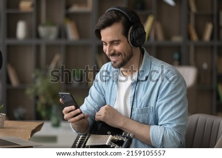 Millennial man musician in headset hold guitar distracted from recording music in audio editing app at home studio on pc to make telephone call. Smiling young guy guitarist talk on phone by video call