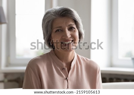 Happy middle aged grey haired lady indoor head shot portrait. Cheerful pretty mature woman wearing casual, looking at camera with toothy smile, posing at home, laughing. Video call screen