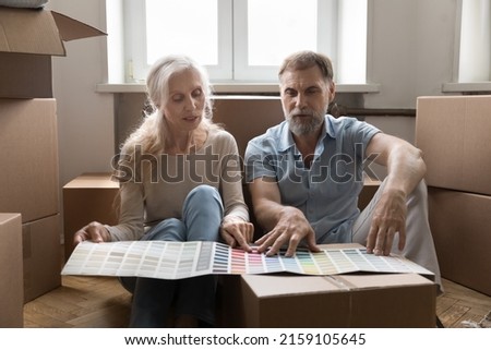 Senior couple discussing wall colors of new home, pointing at swatches in paper brochure on relocation box, planning interior design improvement, decoration, renovation Royalty-Free Stock Photo #2159105645
