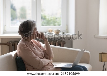 Thoughtful frustrated mature 60s woman holding laptop computer, looking away in deep sad thoughts, thinking over bad concerning news, health problems, sitting on home couch, feeling worried Royalty-Free Stock Photo #2159105699