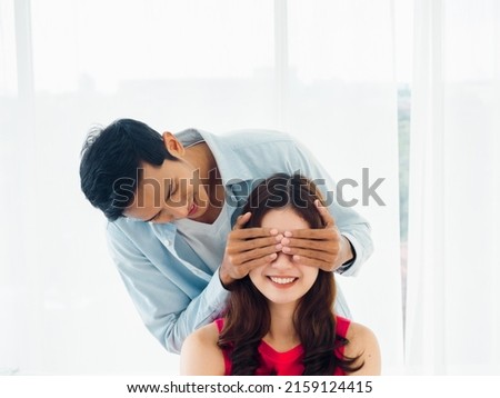 Making surprise, lover, sweet couple concept. Handsome Asian man using hands covering his girlfriend's eyes of excited pretty smiling girl make surprise in bedroom on white curtain background. Royalty-Free Stock Photo #2159124415
