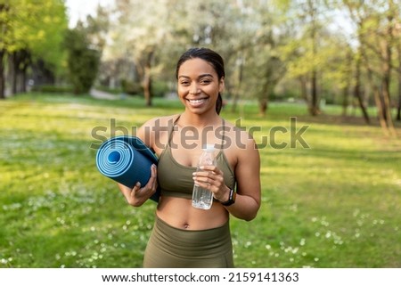 Portrait of cheerful young black woman holding fresh water and sports mat at urban park. Fit African American lady ready for yoga practice, keeping hydrated during outdoor training Royalty-Free Stock Photo #2159141363