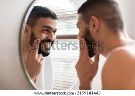 Portrait Of Happy Handsome Arab Man Looking At Reflection In Mirror In Bathroom And Touching Face, Attractive Masculine Middle Eastern Guy Putting Cream On His Cheek. Male Facial Care Concept Royalty-Free Stock Photo #2159141445