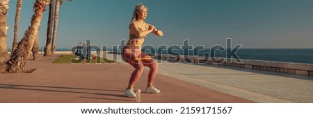On warm sunny day, an active woman performing squats with fitness band on sports street playground near the coast. Royalty-Free Stock Photo #2159171567