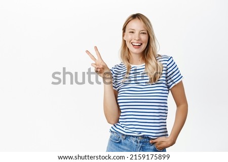 Positive and happy young woman, 25 years old, laughing and smiling, showing v-sign, peace gesture near face, standing over white background