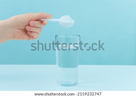 Dry collagen powder in a measuring spoon in hand. A glass of water is on the table.