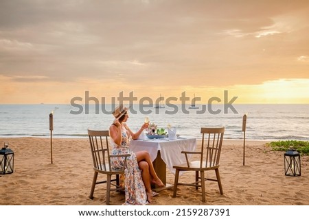 Romantic dinner on sunset. Woman sitting alone on table set with lantern for a romantic meal on beach, yachts and ocean on background. Dinner for a couple in love in luxury outdoor restaurant Royalty-Free Stock Photo #2159287391