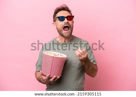 Middle age caucasian man isolated on pink background with 3d glasses and holding a big bucket of popcorns