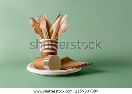 Stack of eco-friendly disposable tableware. Wooden forks and knives, paper cups and plates against green background. Biodegradable cutlery and dishes for picnics, takeaways. Copy space. Front view. Royalty-Free Stock Photo #2159337389