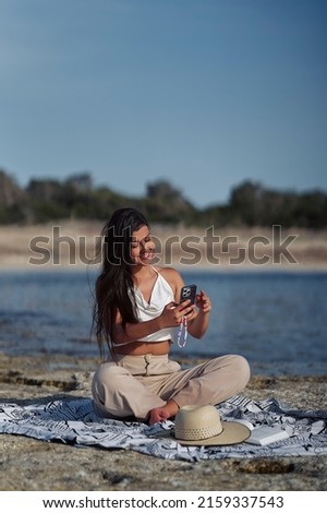 Young latina girl sitting on the beach taking a selfie with her cell phone smiling. Vertical photo.