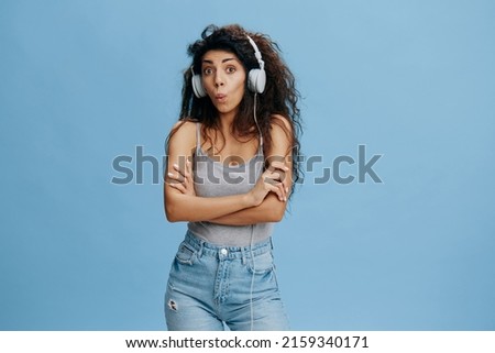 MUSIC STREAMING PLATFORM AD. Shocked awesome curly female in headphones, listen favorite playlist, look at camera, cross hands on chest, say Wow not bad. Studio shoot isolated blue sky background
