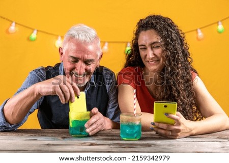 father and daughter have fun putting a smartphone into a glass full of a blue cocktail - studio shot