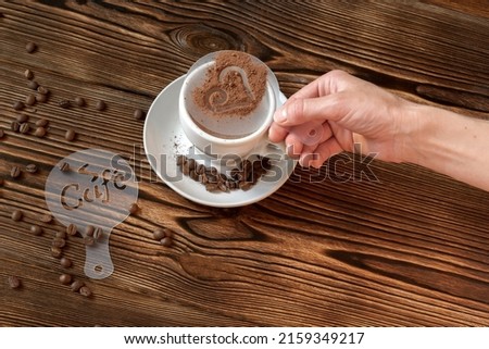 Top view of ceramic cup of hot cappuccino coffee latte with drawing picture of heart shape with cinnamon or cocoa on milk foam, woman hands holding different stencils, coffee beans on wooden table.
