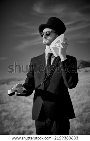 A serious man in a black suit, top hat and black sunglasses stands in a field and calls on the phone, holding a telephone receiver and a telephone set in reverse. Black-and-white photo. Surrealism.