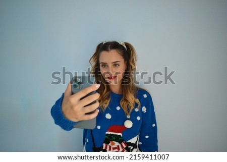 A young adult woman wearing a funny winter sweater and taking a selfie against a gray wall