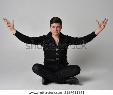 Full length portrait of  handsome brunette male model wearing black shirt and  elegant vest. sitting  Pose with gestural hands reaching out,  isolated on studio background.