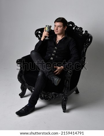 Full length portrait of  handsome brunette male model wearing black shirt and elegant vest. Sitting pose in gothic armchair, with gestural hands,  isolated on studio background.