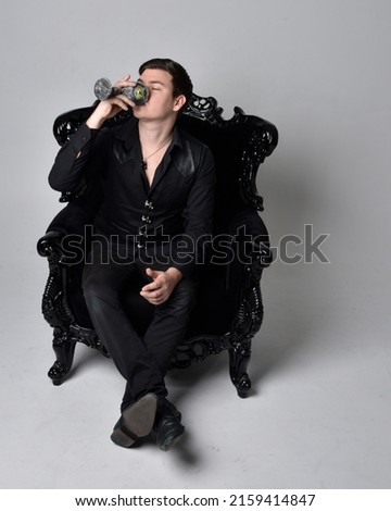 Full length portrait of  handsome brunette male model wearing black shirt and elegant vest. Sitting pose in gothic armchair, with gestural hands,  isolated on studio background.