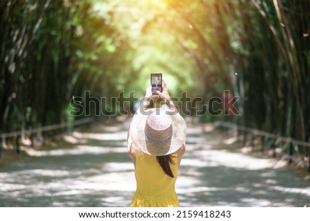 Asian Woman in yellow dress and hat Traveling at green Bamboo Tunnel, Happy traveler taking photo by mobile phone at Chulabhorn wanaram temple in Nakhon Nayok, Thailand