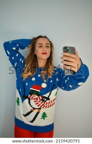 A young adult woman wearing a funny winter sweater and taking a selfie against a gray wall