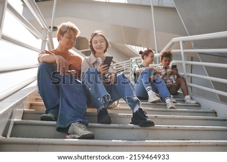 Sunlit full length shot of diverse teenagers using smartphones outdoors while sitting on metal stairs Royalty-Free Stock Photo #2159464933