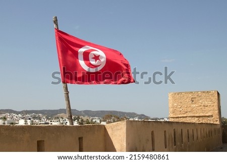 Tunisian National Flag flying from the Old Medina in Hammammet. The crescent and star depicted on the flag of Tunisia are traditional symbols of Islam, and are also considered symbols of good luck. Royalty-Free Stock Photo #2159480861