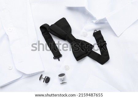 Stylish black bow tie and cufflinks on white shirt, top view