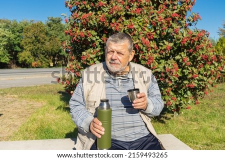 Portrait of ukrainian senior man sitting on  bench against rowan tree and holding vacuum bottle getting ready to drink tea at sunny autumnal day Royalty-Free Stock Photo #2159493265