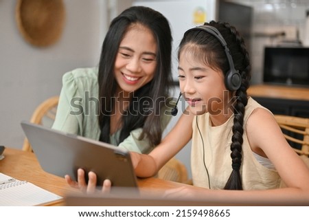 Cute Asian girl in casual clothes and headphones using digital tablet, listening music, watching funny cartoon with her mom in the living room.