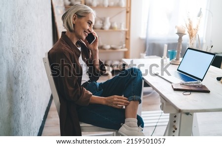 Smiling senior caucasian mature businesswoman sitting on chair near table and talking on smartphone. Concept of small business and entrepreneurship. Modern successful woman. Home art studio. Side view