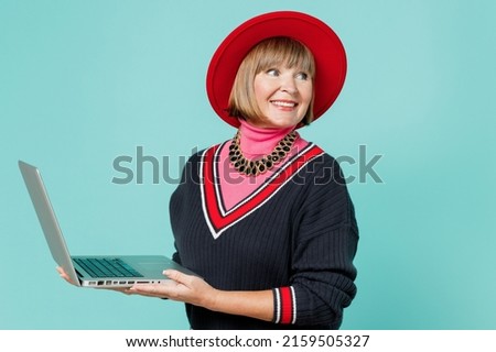 Elderly smiling happy businesslady woman 50s in necklace shirt red hat hold use work on laptop pc computer look aside on copy space area isolated on plain pastel light blue background studio portrait