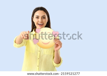Beautiful young woman holding smiling emoticon with hearts on light blue background